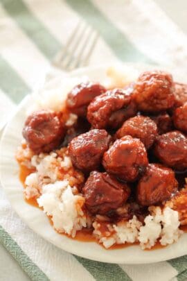 A plate of white rice topped with glazed meatballs, placed on a green and white striped cloth with a fork in the background.