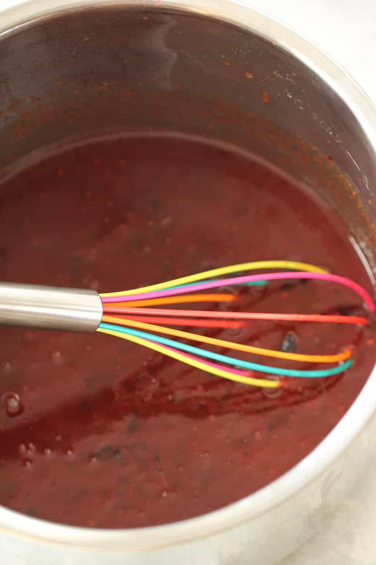 A saucepan filled with dark red liquid is being stirred by a rainbow-colored whisk.