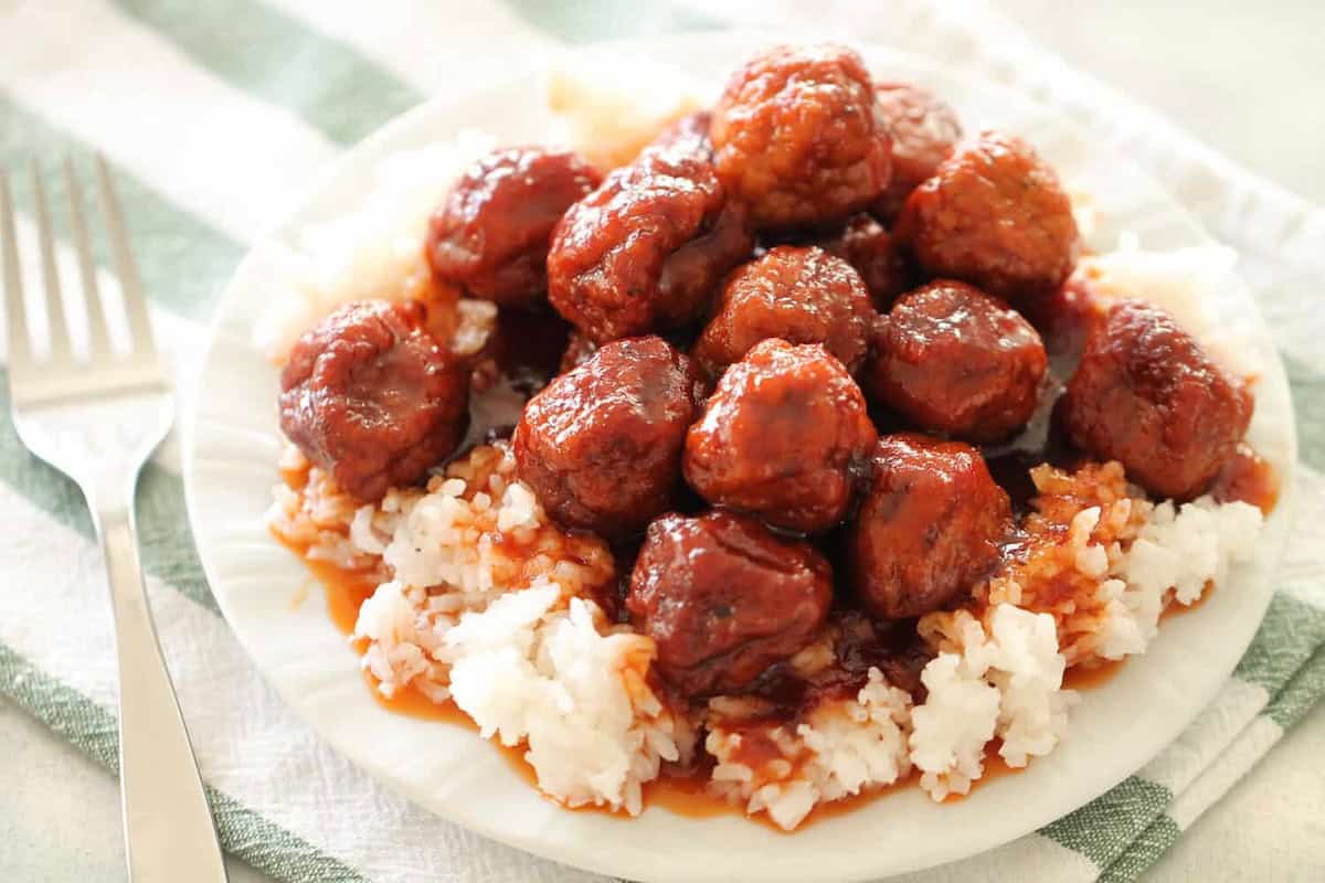 Plate of glazed meatballs on white rice with a fork on the side, set on a striped cloth.