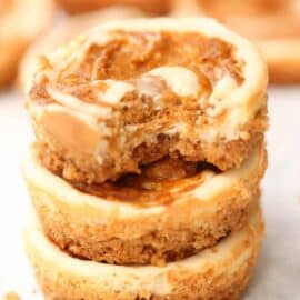 Stack of three mini cheesecakes with a swirl of caramel on top. A bite is taken out of the top cheesecake, revealing a creamy interior and crumbly crust.