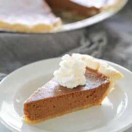 Slice of pumpkin pie with a dollop of whipped cream on top, served on a white plate, with the remaining pie in the background.