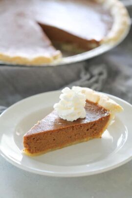 Slice of pumpkin pie with a dollop of whipped cream on top, served on a white plate, with the remaining pie in the background.