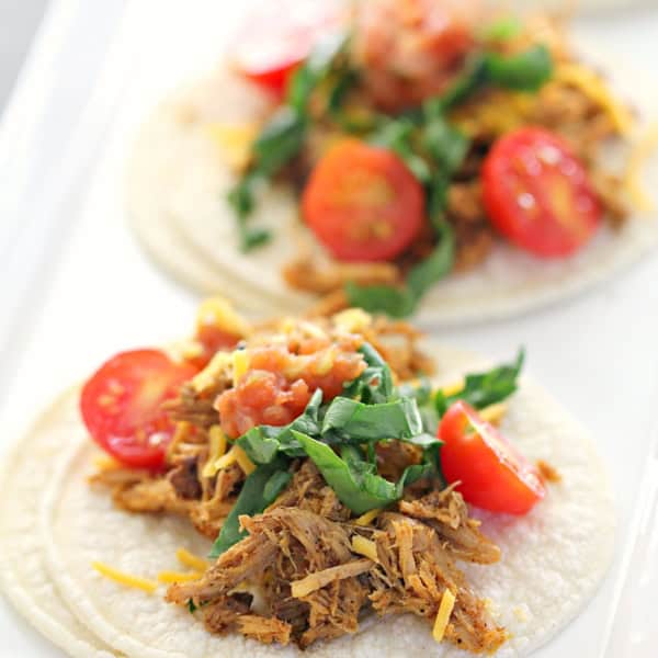 A plate with three soft tortillas topped with shredded meat, chopped greens, cherry tomatoes, and a sprinkle of cheese.