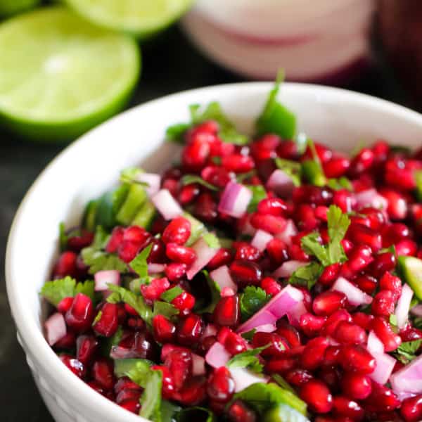 A bowl of pomegranate salsa with chopped red onions and cilantro, garnished with lime slices in the background.