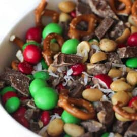 A bowl of snack mix with red and green candies, pretzels, peanuts, chocolate chunks, and shredded coconut.