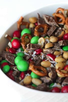 A bowl of snack mix with red and green candies, pretzels, peanuts, chocolate chunks, and shredded coconut.