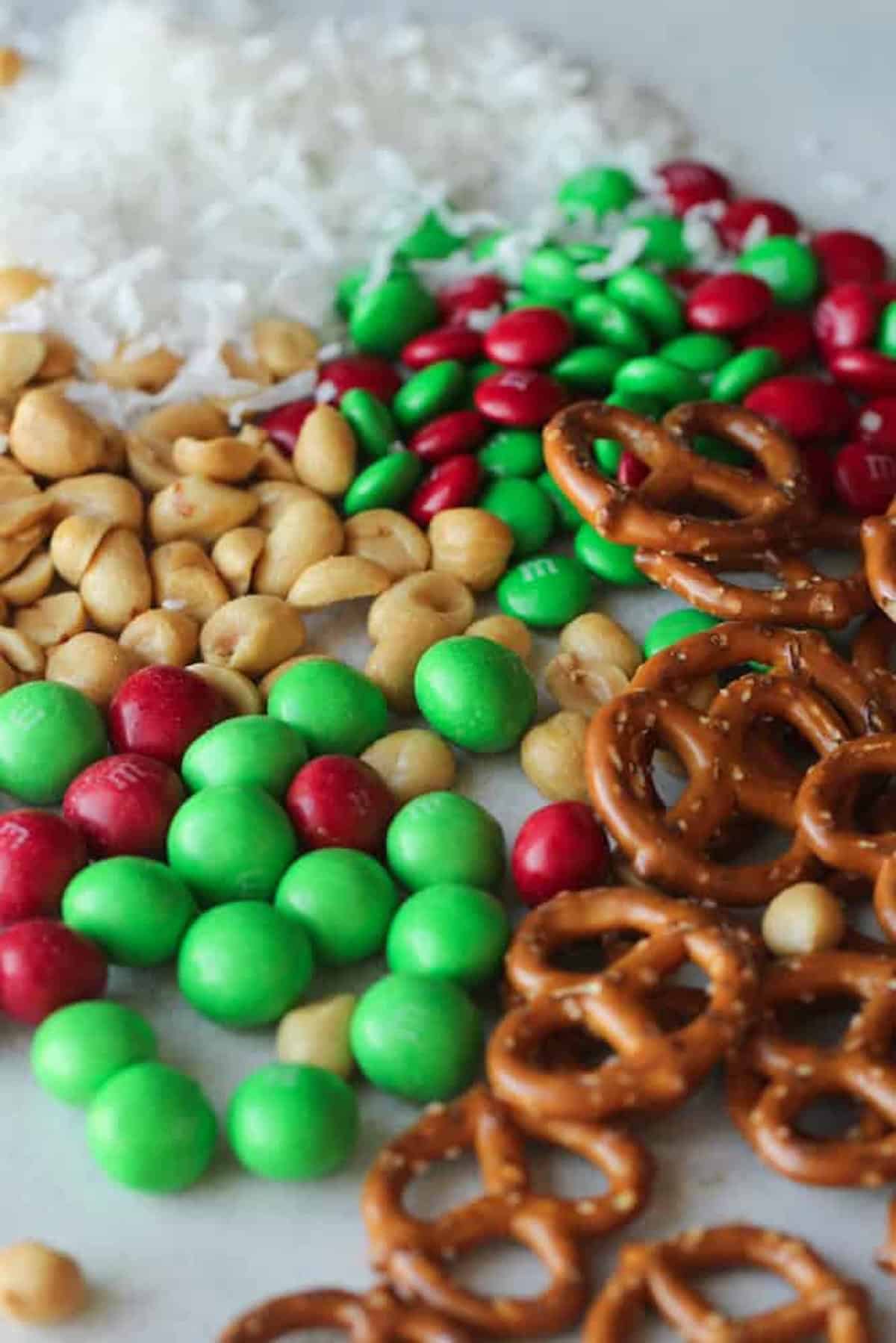 Assorted snacks on a surface: red and green candy-coated chocolates, pretzels, peanuts, and shredded coconut.