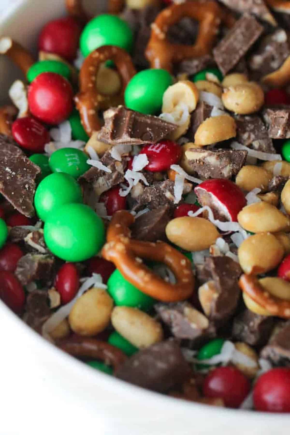 A bowl filled with pretzels, red and green candies, peanuts, chocolate chunks, and coconut flakes.