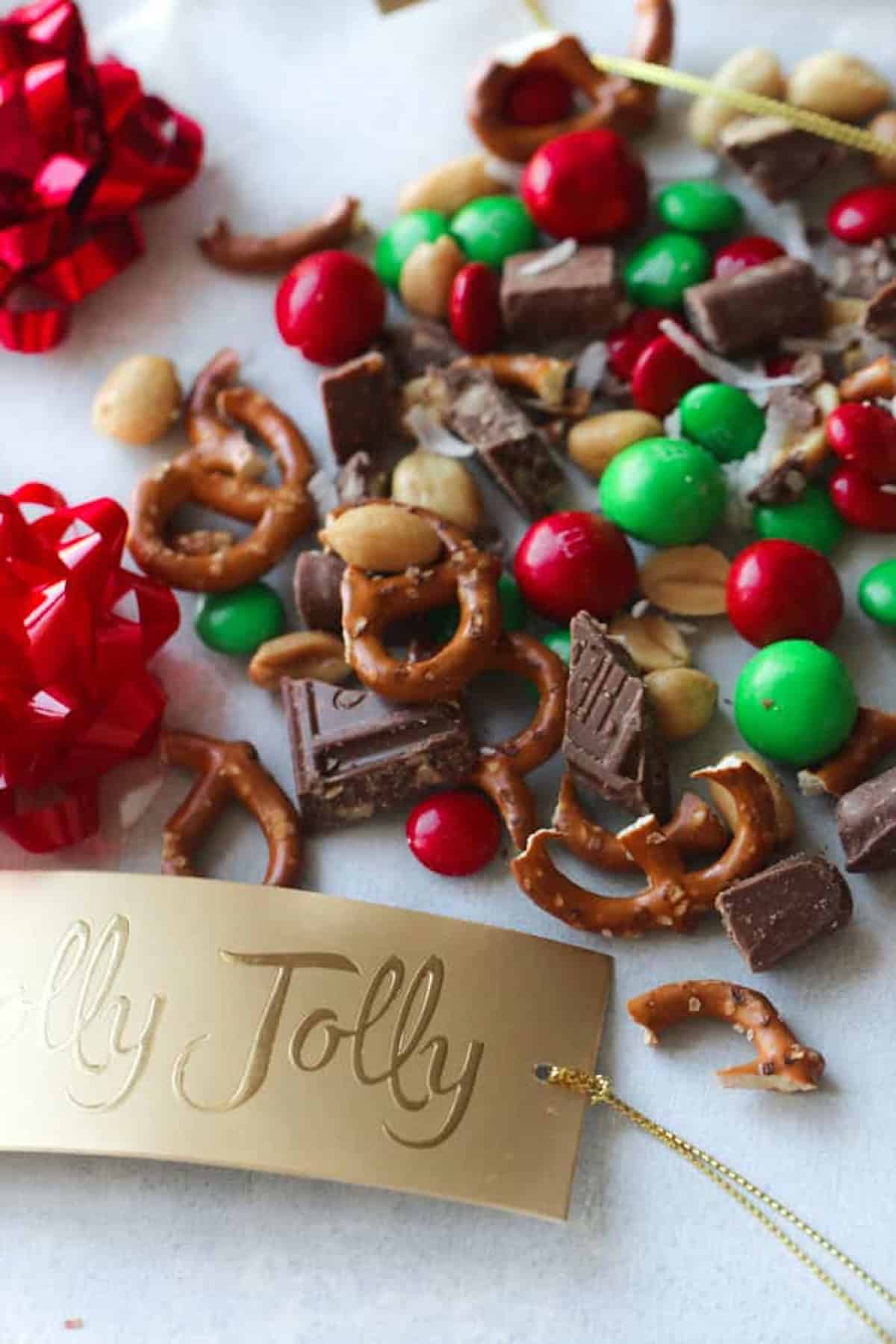 A festive mix of pretzels, peanuts, chocolate pieces, and red and green candy with red bows and a "Jolly Jolly" tag on a white surface.