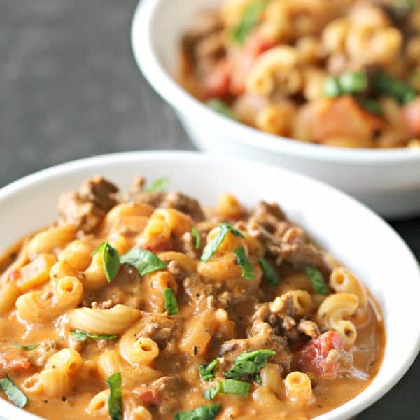 Two white bowls filled with macaroni beef stew, garnished with chopped herbs, on a dark surface.