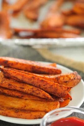 A plate of seasoned sweet potato fries with a small bowl of red dipping sauce in the foreground.