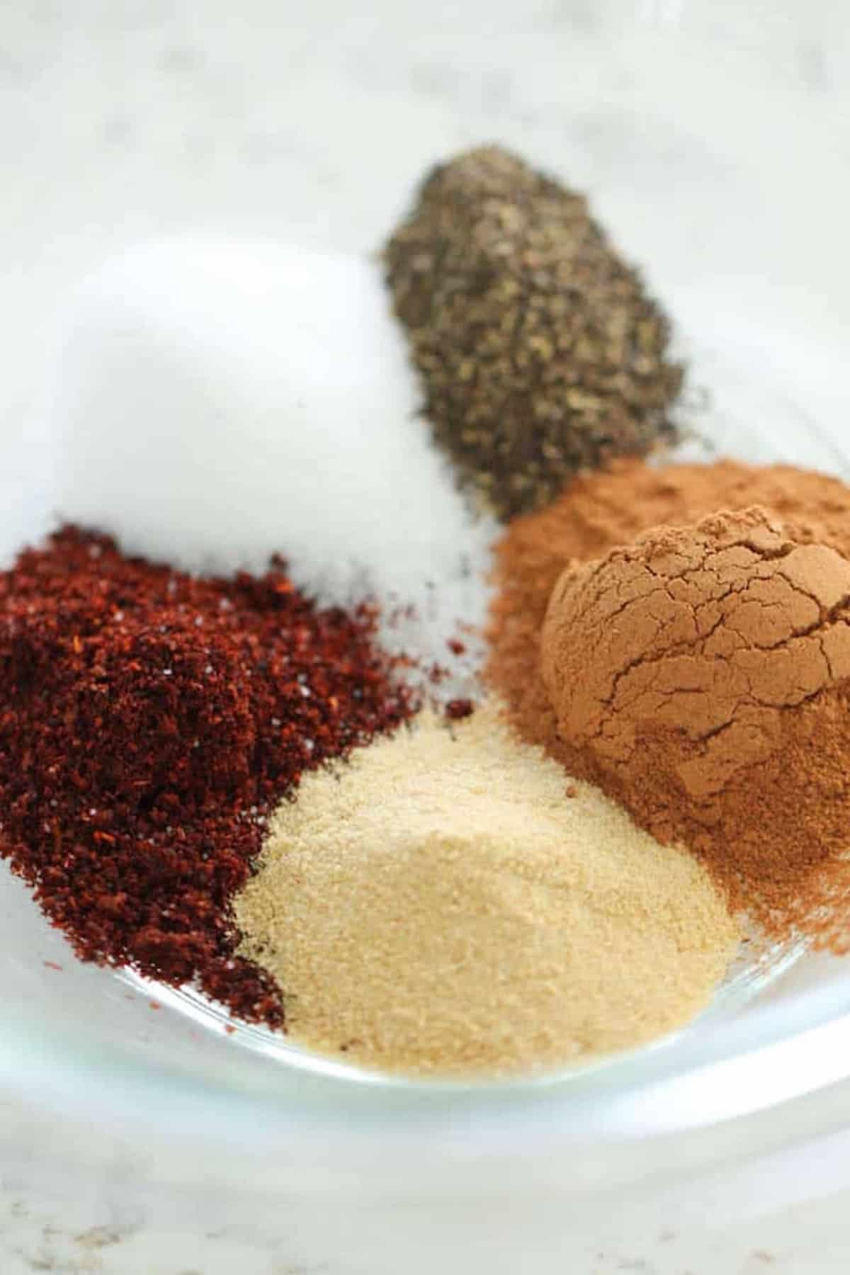 A small glass bowl containing five different ground spices, including black pepper, salt, chili powder, cinnamon, and garlic powder, arranged in sections.