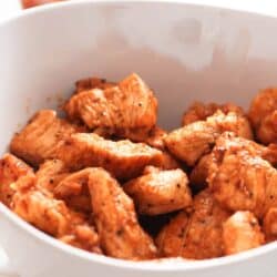 A bowl of seasoned, cooked chicken pieces, with a bottle of hot sauce in the background.