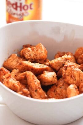 A bowl of seasoned, cooked chicken pieces, with a bottle of hot sauce in the background.