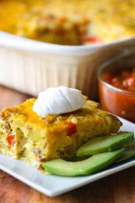 Close-up of a slice of savory casserole topped with sour cream on a plate, accompanied by avocado slices. A dish of salsa is visible in the background.