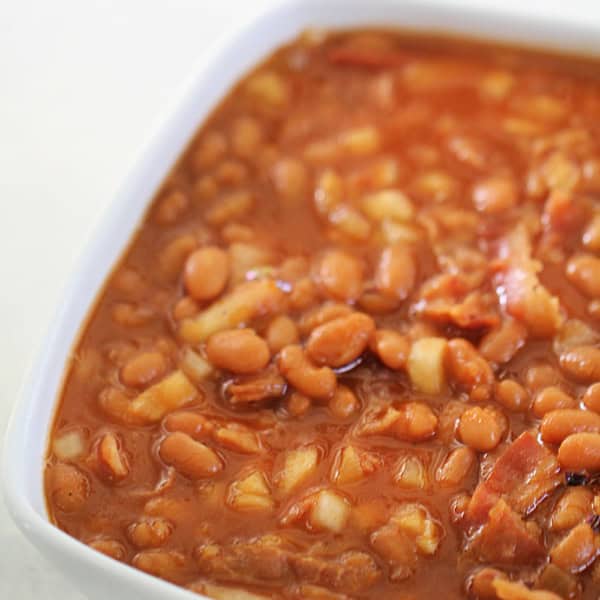 A square white dish filled with baked beans in a rich, reddish-brown sauce with visible chunks of onion and bacon.
