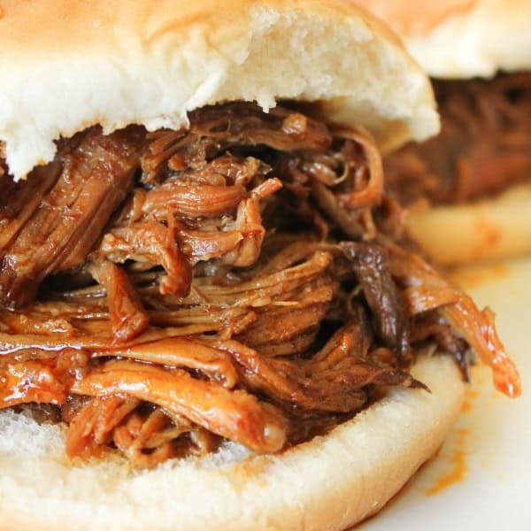 Close-up of a sandwich filled with smoky BBQ pulled pork on a soft bun, with another similar sandwich partially visible in the background.