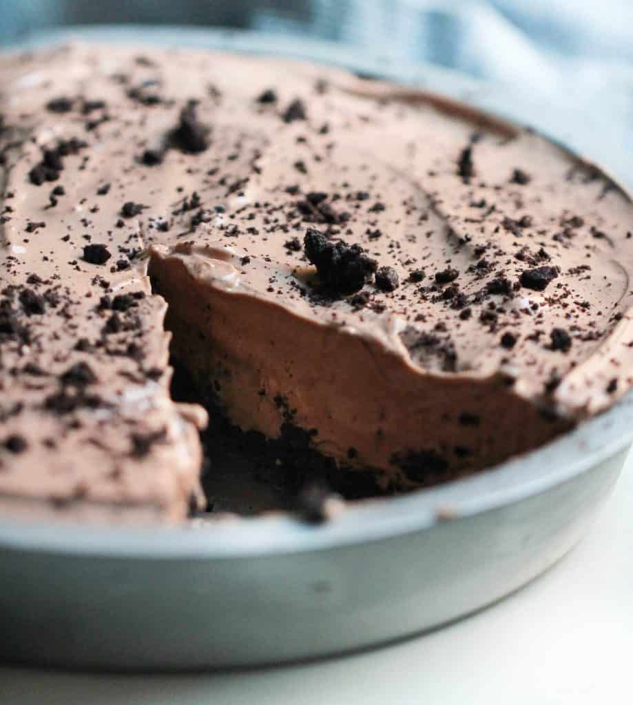 A close-up of a chocolate oreo pie topped with crumbled bits in a round metal pan on a light background with a slice missing