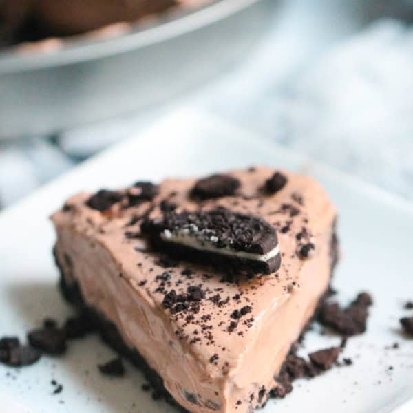 A slice of chocolate cream pie topped with crushed cookies and a large cookie piece on a square white plate, with the whole pie visible in the background.