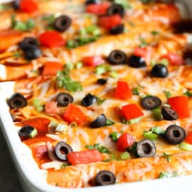 A close-up of a casserole dish filled with enchiladas, topped with melted cheese, sliced black olives, diced red bell peppers, and chopped green onions.