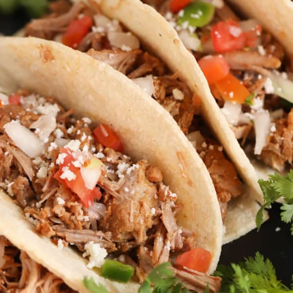 Three soft shell tacos filled with shredded meat, diced tomatoes, onions, green peppers, and topped with crumbled cheese, garnished with fresh cilantro.