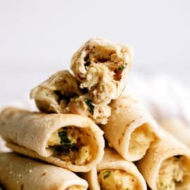 A stack of rolled tortillas filled with a mixture of scrambled eggs, herbs, and likely meat, with one tortilla on top broken in half to display the filling.