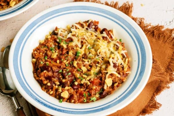 A bowl of Mom's Slow Cooker Chili topped with shredded cheese and chopped green herbs, placed on a brown napkin with a spoon beside it. Another partially visible bowl and a small dish of herbs are in the background.