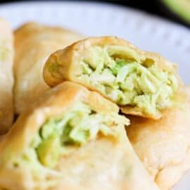 Close-up of golden-brown pastries filled with a green avocado mixture on a white plate. One pastry is cut open to show the filling.