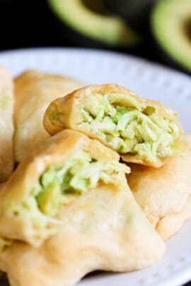 Close-up of golden-brown pastries filled with a green avocado mixture on a white plate. One pastry is cut open to show the filling.