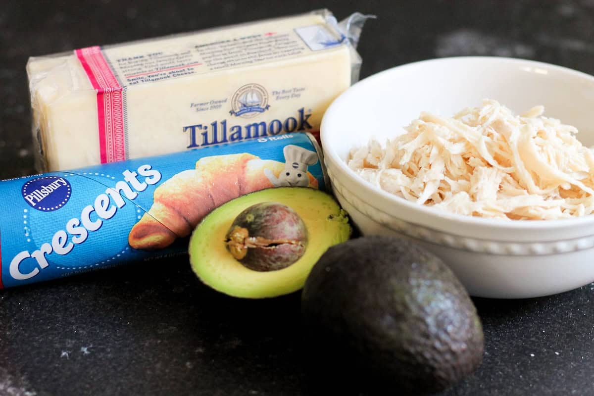 A block of Tillamook cheese, a can of Pillsbury Crescents, a halved avocado, a whole avocado, and a bowl of shredded chicken on a black surface.