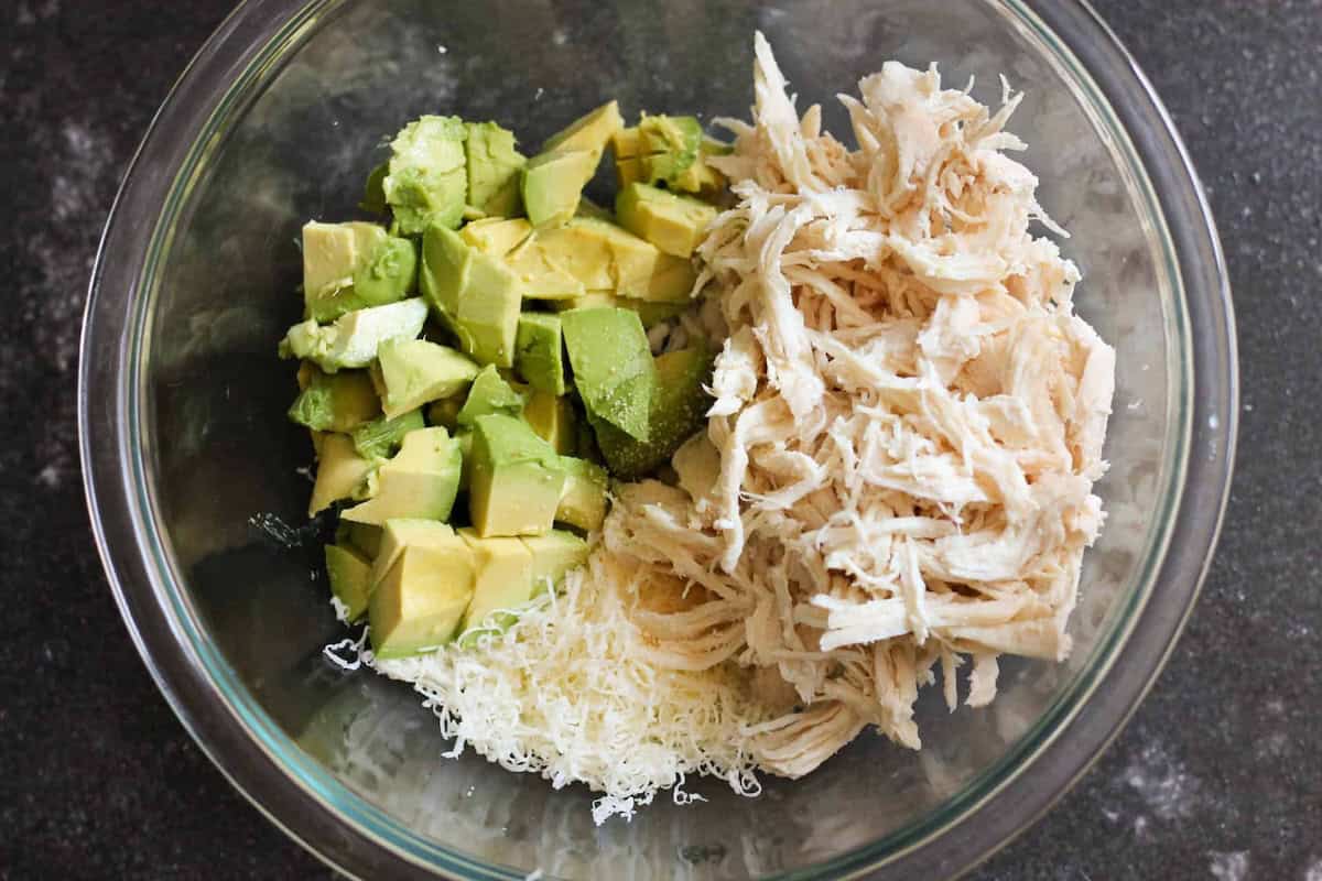 A glass bowl containing cubed avocado, shredded chicken, and grated cheese on a dark surface.