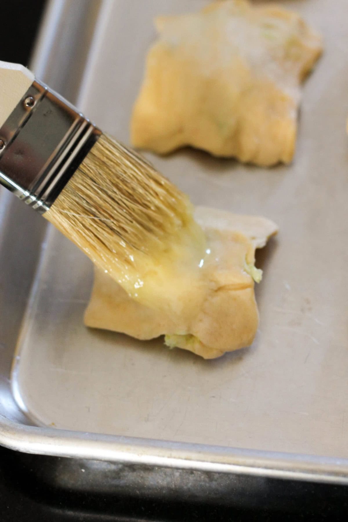 A pastry brush is applying an egg wash to a piece of pastry dough on a baking tray.