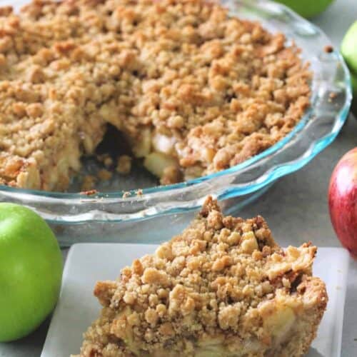 A pie with a crumbly topping is in a glass dish. A slice has been cut out and placed on a white plate in the foreground. Several whole apples are placed around the pie.