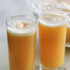 Two tall glasses filled with a frothy orange juice sit on a light-colored surface. A large jug of the same juice is partially visible in the background.