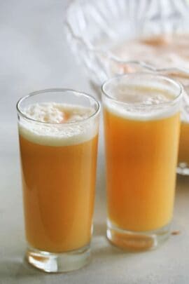 Two tall glasses filled with a frothy orange juice sit on a light-colored surface. A large jug of the same juice is partially visible in the background.