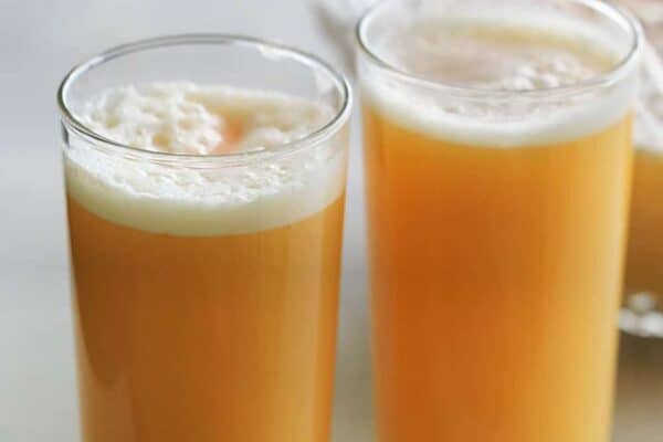 Two tall glasses filled with a frothy orange juice sit on a light-colored surface. A large jug of the same juice is partially visible in the background.