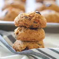 A stack of three chocolate chip cookies sits on a striped cloth, with more cookies on a baking tray in the background.