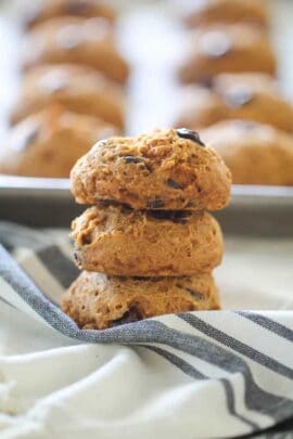 A stack of three chocolate chip cookies sits on a striped cloth, with more cookies on a baking tray in the background.