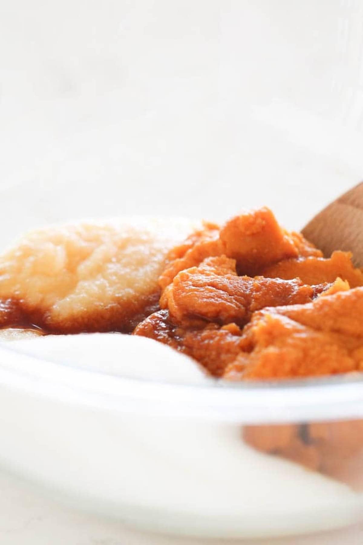 A clear glass bowl containing mashed pumpkins and applesauce, with a wooden spoon partially visible.