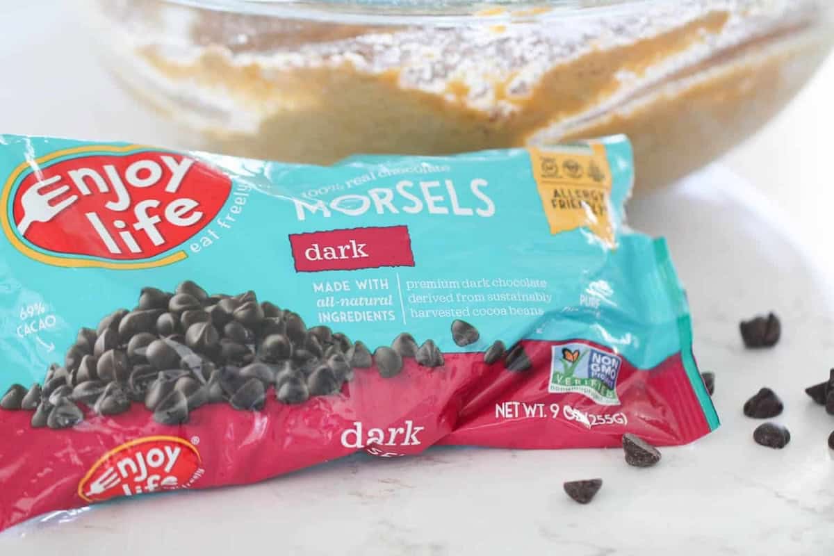 A bag of Enjoy Life dark chocolate morsels is placed on a counter with some spilled morsels next to it. A mixing bowl with batter is visible in the background.