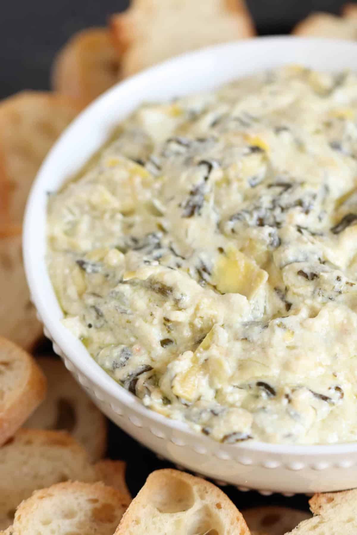 A bowl of creamy spinach and artichoke dip surrounded by slices of toasted baguette.