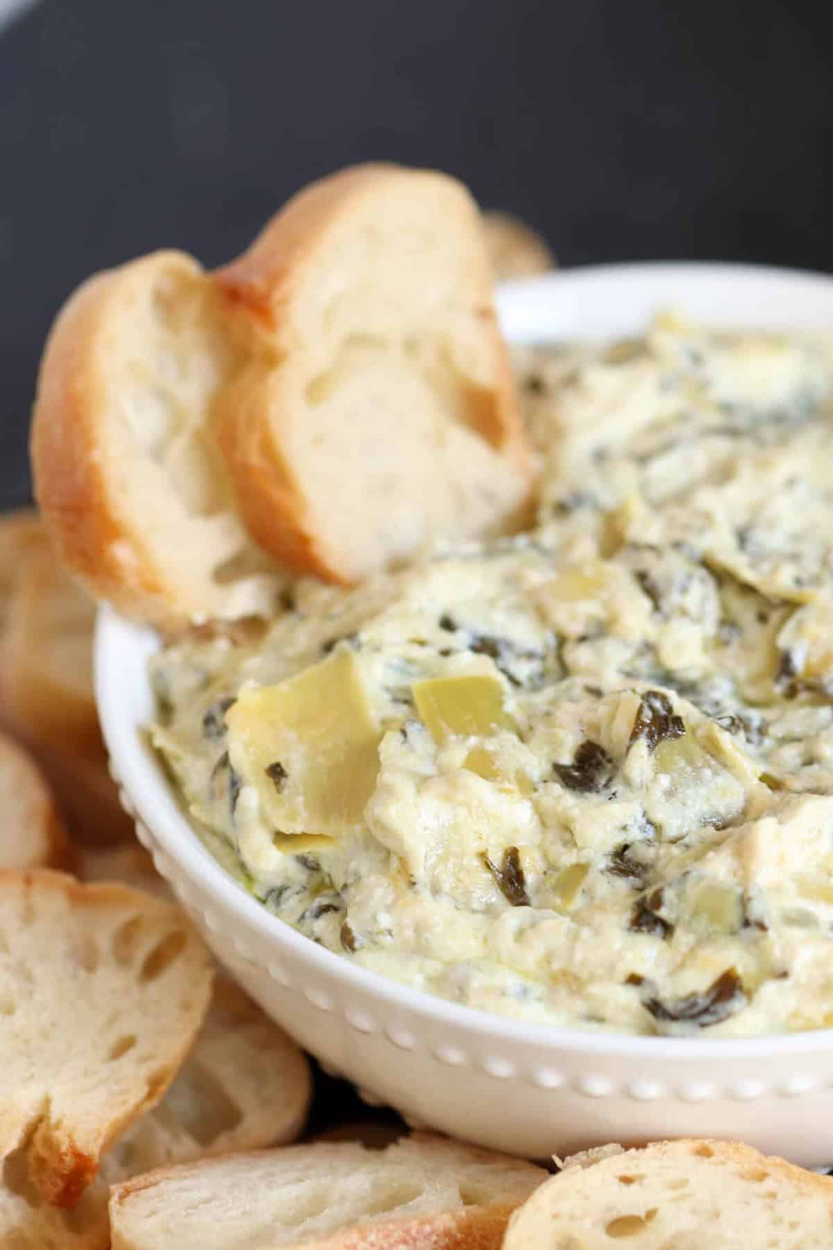 A bowl of creamy spinach and artichoke dip surrounded by sliced baguette pieces.