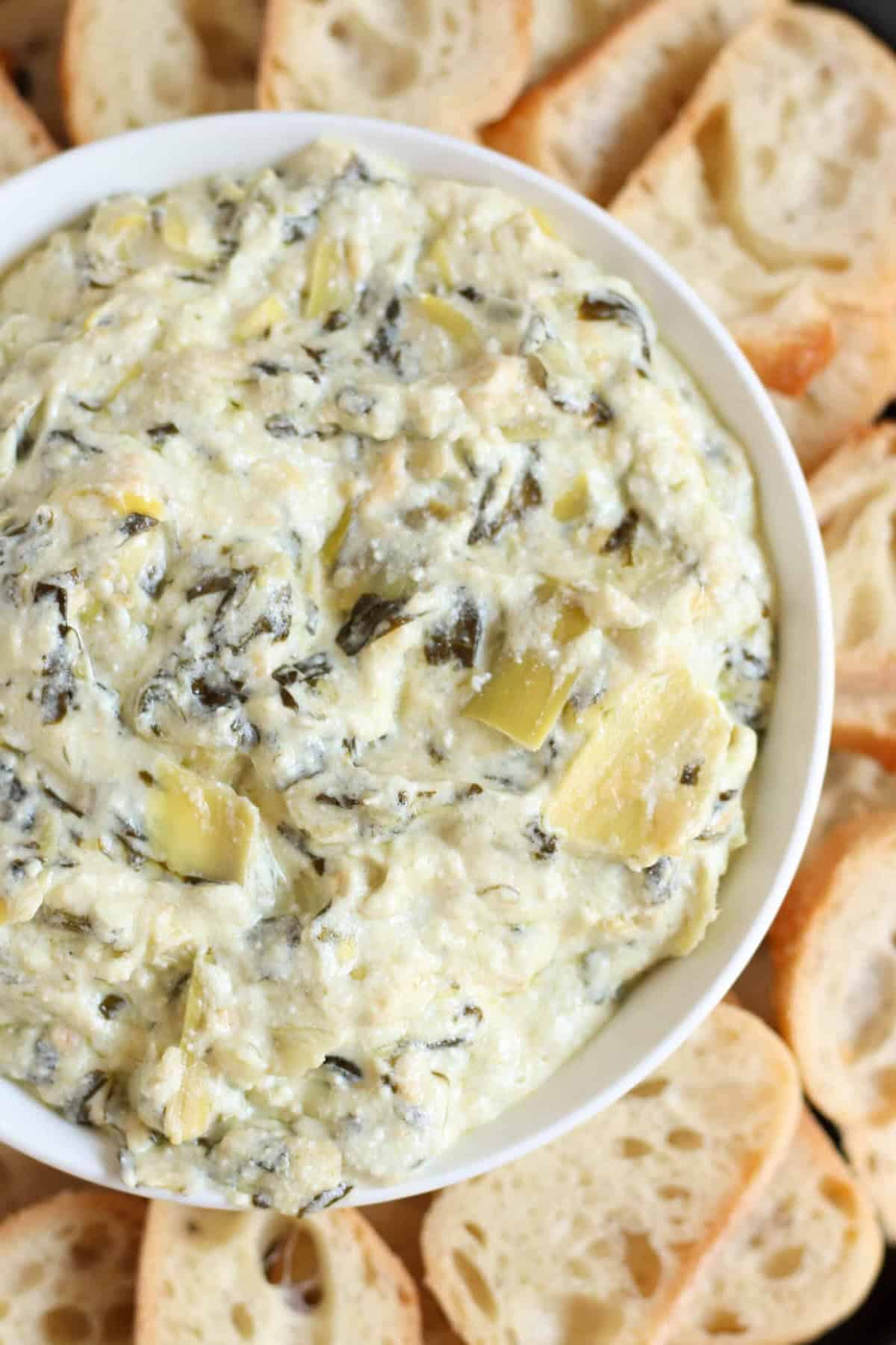 A bowl of creamy artichoke dip surrounded by sliced baguette pieces.