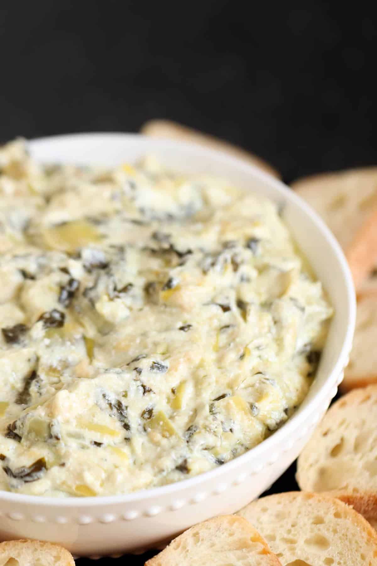 A bowl of creamy spinach and artichoke dip surrounded by sliced bread pieces on a black background.