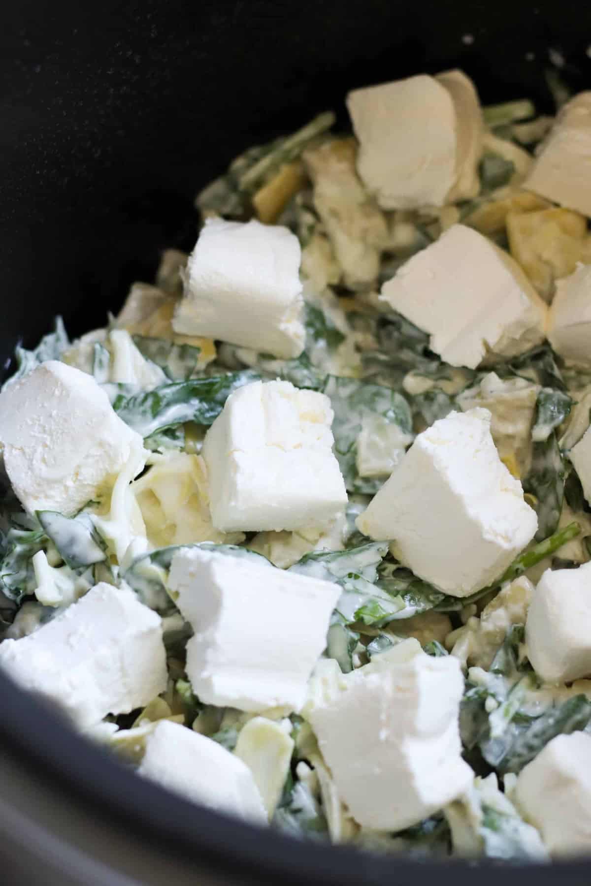 Close-up of a dish containing chopped spinach, artichoke, and cream cheese cubes, all mixed with a creamy sauce.