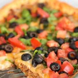 Close-up of a freshly baked pizza topped with diced tomatoes, black olives, chopped greens, and melted cheese in a glass baking dish. A metal spatula is partly visible underneath the pizza.