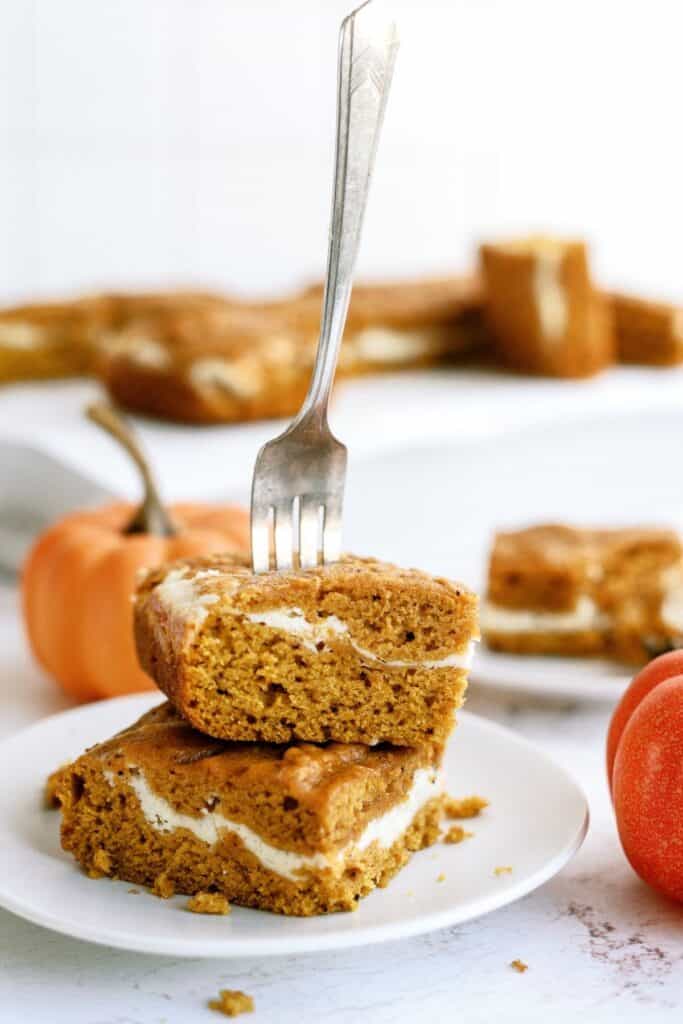 A fork pierces two pumpkin bars with a cream cheese filling, stacked on a white plate. Small pumpkins can be seen in the background.