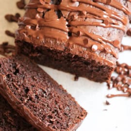 A sliced loaf of chocolate bread, topped with a drizzle of chocolate frosting, is displayed on a light-colored surface.