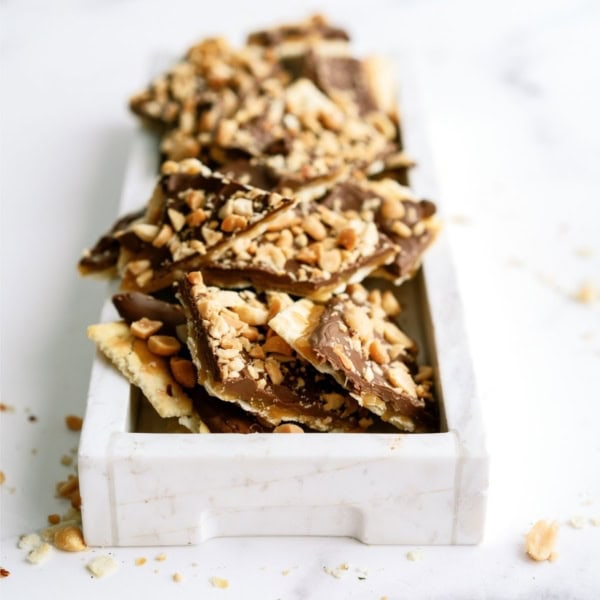 A white tray filled with irregular pieces of chocolate-covered toffee bark, garnished with chopped nuts.