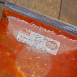 Close-up of a plastic zipper bag filled with red soup labeled \"Taco Soup\" and instructions written on the label. The bag is on a wooden surface.